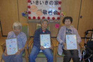 入居者による敬老会での作品展示。3名が絵画作品を掲げて記念撮影。